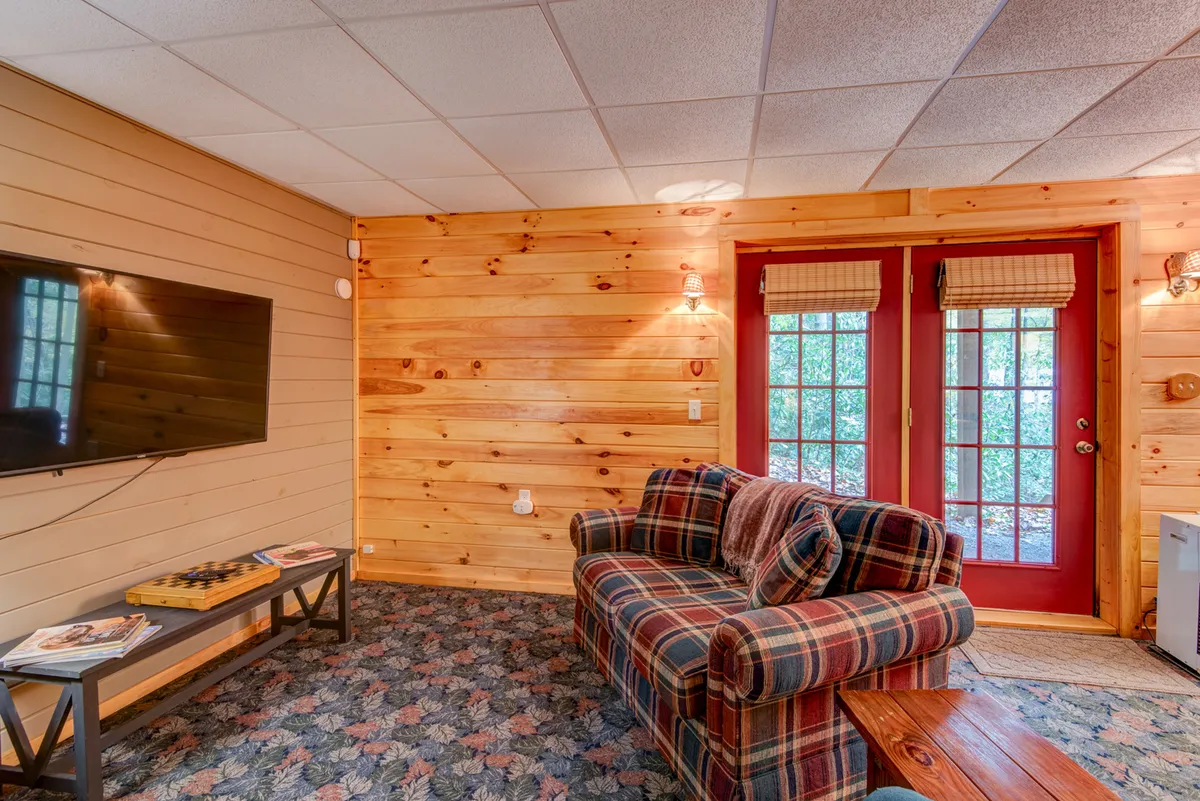 A cozy entertainment area with a plaid couch, double doors, television, and a wooden coffee table. The room is designed for relaxing, enjoying movies, or gathering for board games, while the doors subtly hint at access to the outdoors.