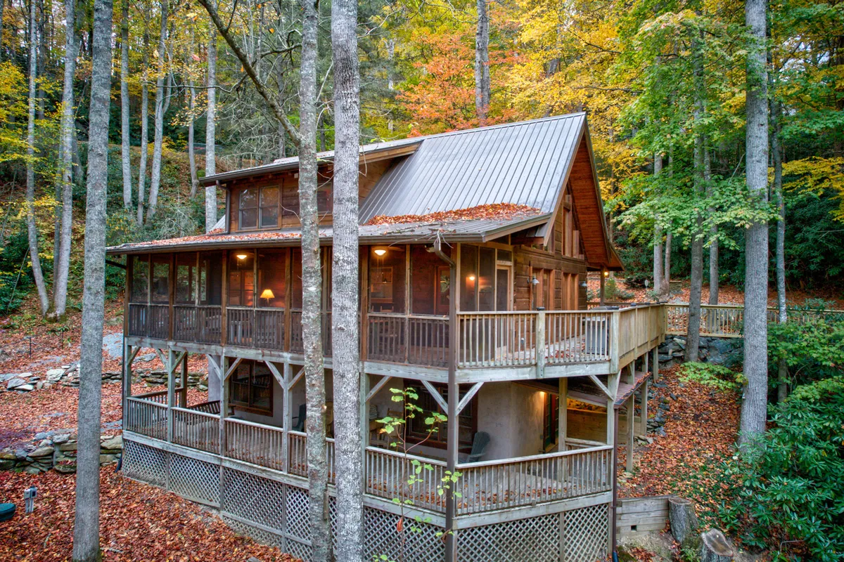 A cozy wooden cabin nestled amongst tall trees, with a spacious decking area surrounded by vibrant autumn leaves. The outdoor space is inviting, featuring areas perfect for gatherings and open nature trails extending into the woods.
