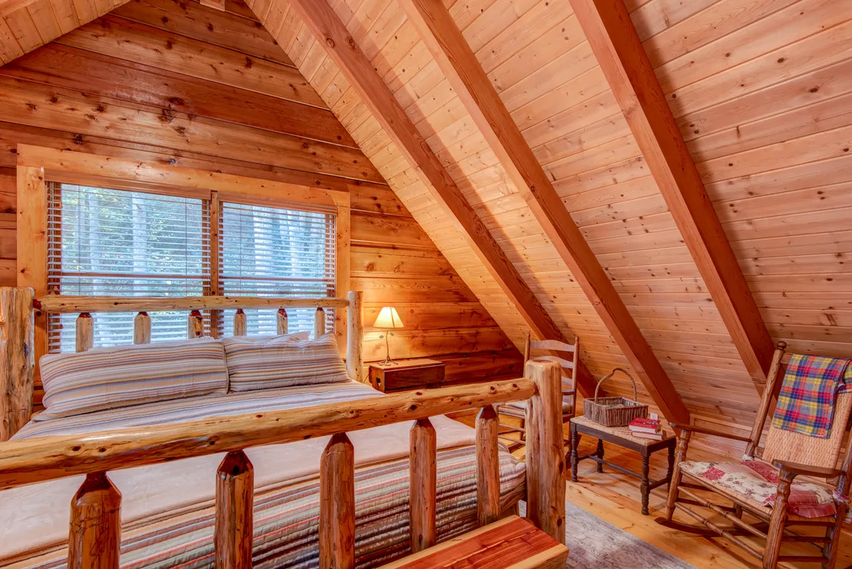 A warmly lit bedroom with a wooden bed frame, striped bedding, angled wooden beams in the ceiling, a cozy table lamp, and a wooden rocking chair. The window with blinds allows natural light to filter in, creating a peaceful, inviting retreat.