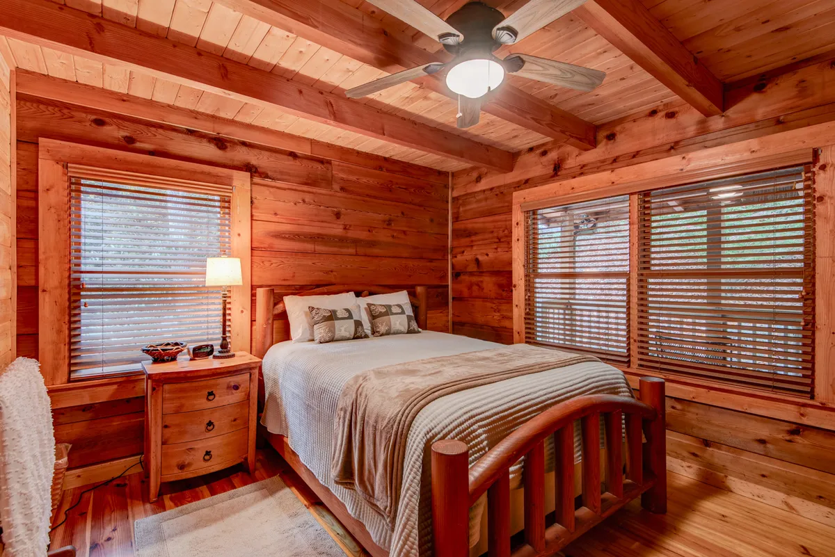 A comfortable queen bedroom with a wooden bed, nightstand with lamp, and windows letting in soft daylight. The room is intimate and peaceful, offering a restful place to recharge.