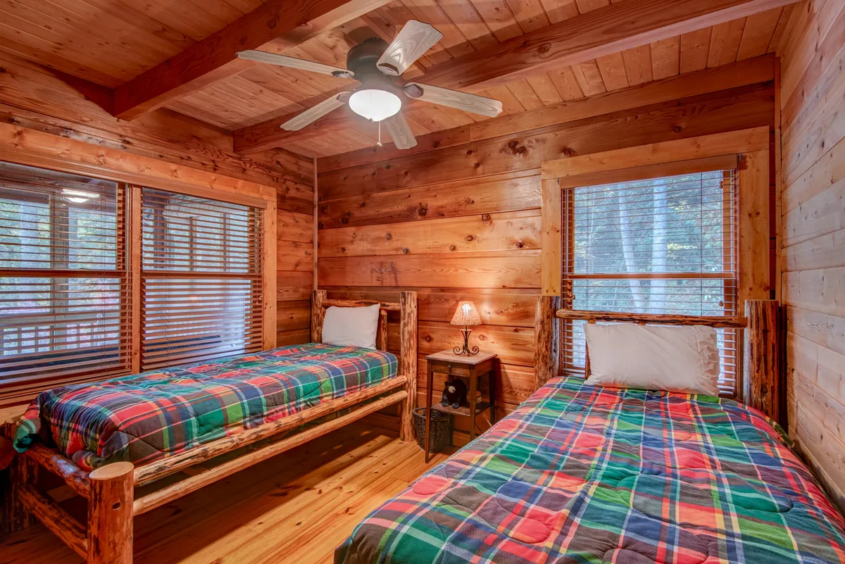 A kid-friendly bedroom featuring two wooden beds with plaid bedding, wooden walls, windows with blinds, and a nightstand with a lamp. The ambiance is warm and homey, ideal for children or extra guests.