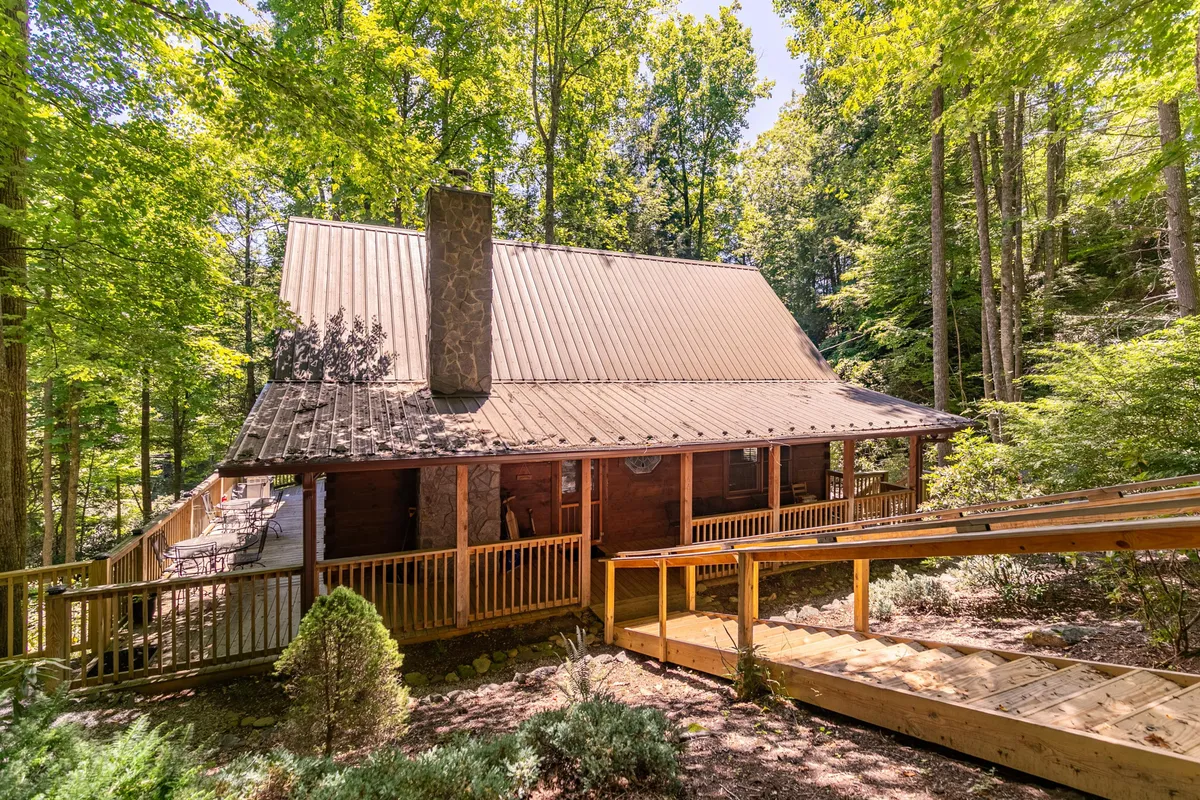 This image features the cabin's striking exterior, with its classic wooden structure, a spacious wooden deck, a prominent stone chimney, and lush trees all around. The peaceful setting suggests both relaxation and a connection to nature, perfect for unwinding after outdoor adventures. The deck offers space for gatherings or quiet moments to take in the surroundings.