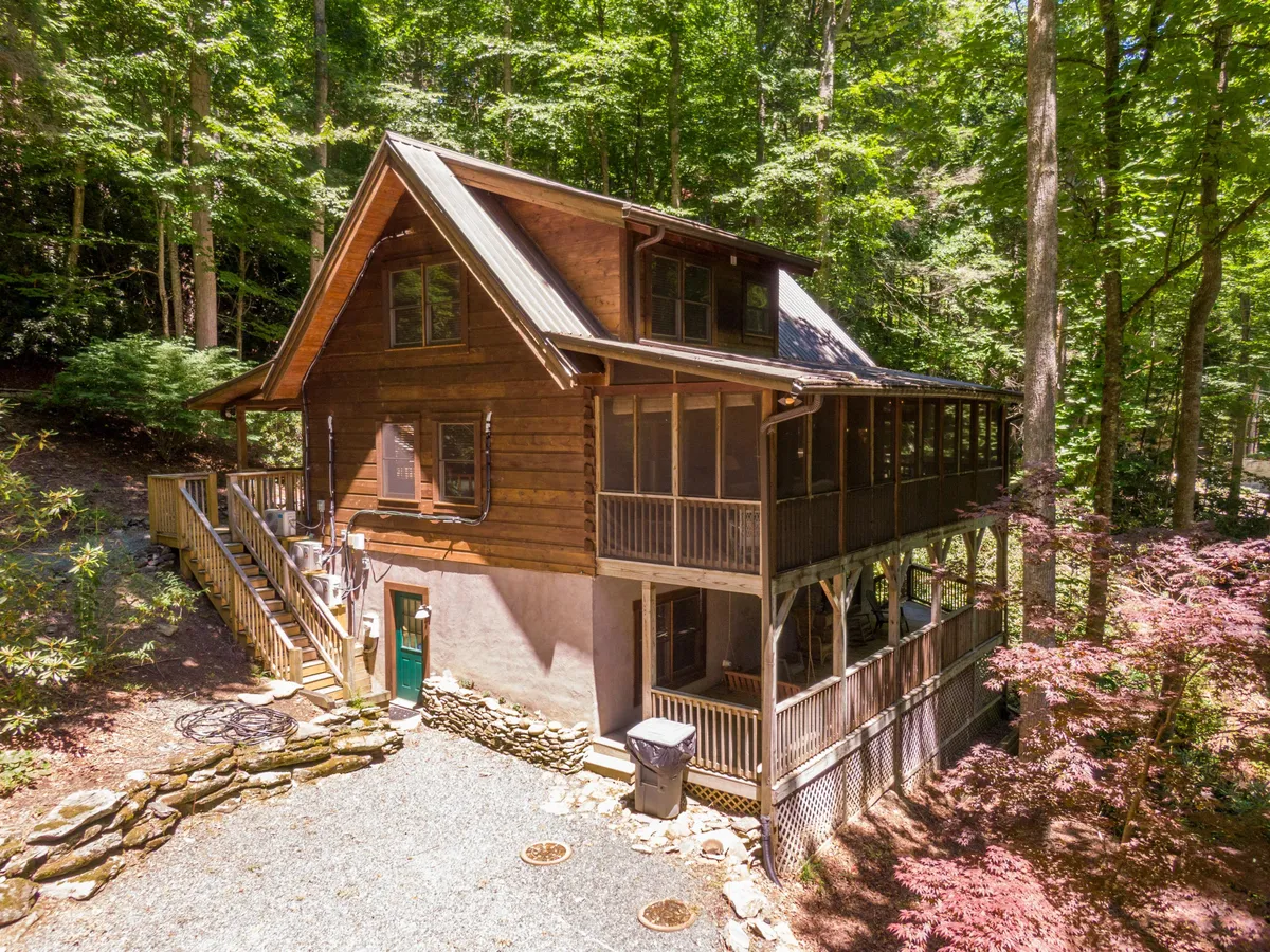 This image highlights the wooden cabin structure with a prominent wrap-around porch and a screened-in porch area, set amidst tall trees and a serene natural backdrop. The stone pathway leads up to the porch, inviting guests to imagine themselves enjoying leisurely mornings or evenings outdoors. The ambiance is tranquil and cozy, making this a perfect visual for outdoor living.