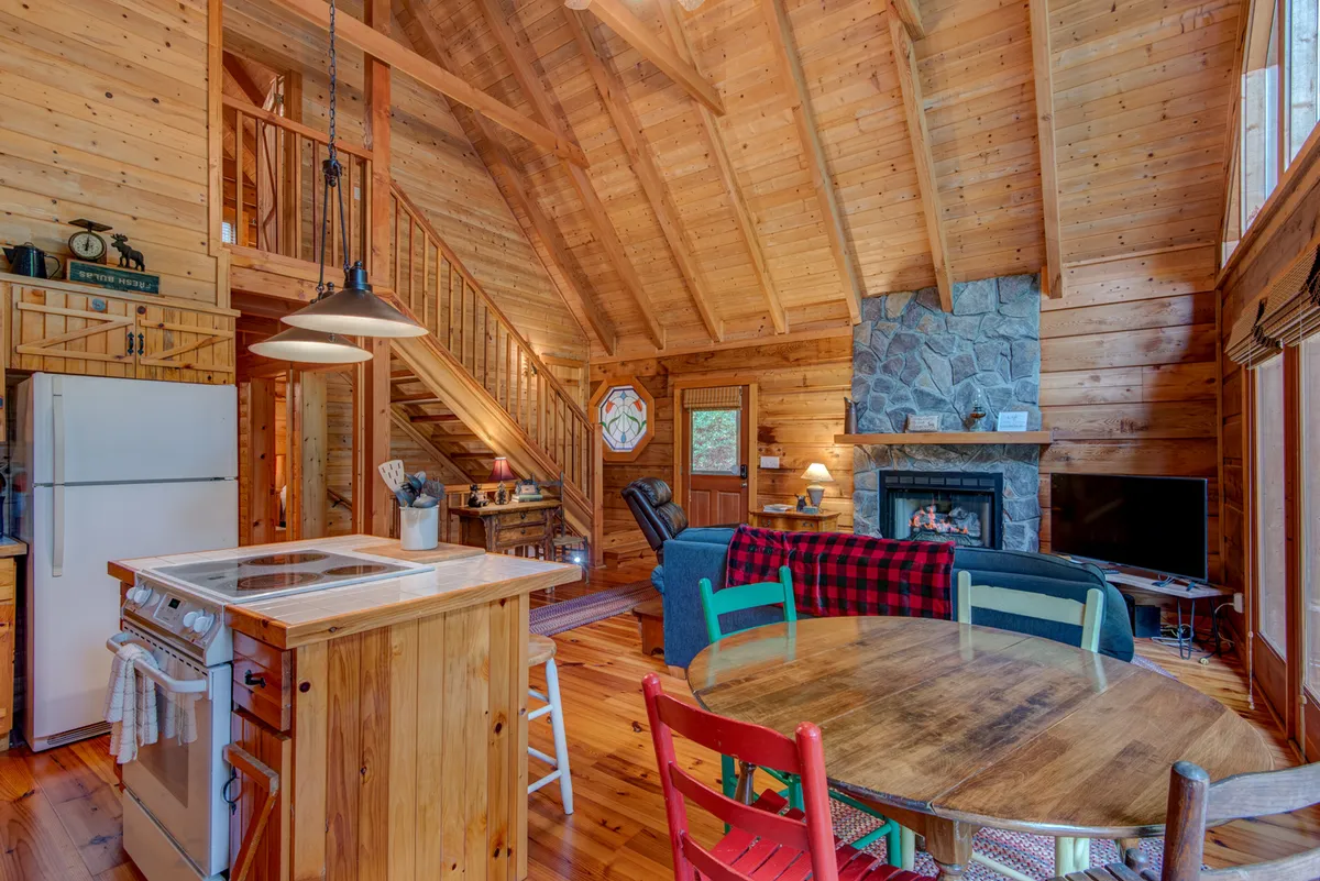 A warm, inviting interior where the dining area with colorful chairs sits adjacent to a stone fireplace, with the kitchen island nearby. Exposed beams and wood finishes create a rustic, cohesive look ideal for family gatherings and shared meals.