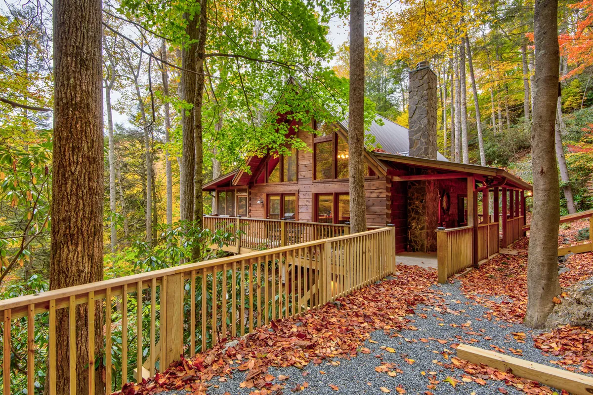 A cozy log cabin surrounded by towering trees, featuring a classic stone chimney and inviting wooden railing. The warm glow of sunset filters through the forest, hinting at peaceful evenings and a tranquil mountain retreat.