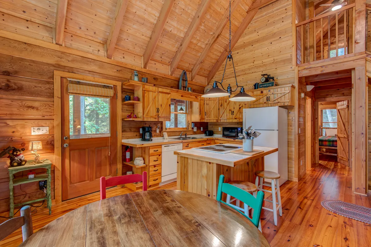 An open-concept kitchen and dining area featuring a central island, wooden beams overhead, and a dining table with chairs beside a large window. Natural light pours in, illuminating the space and offering scenic views, while the kitchen is ready for preparing family meals.