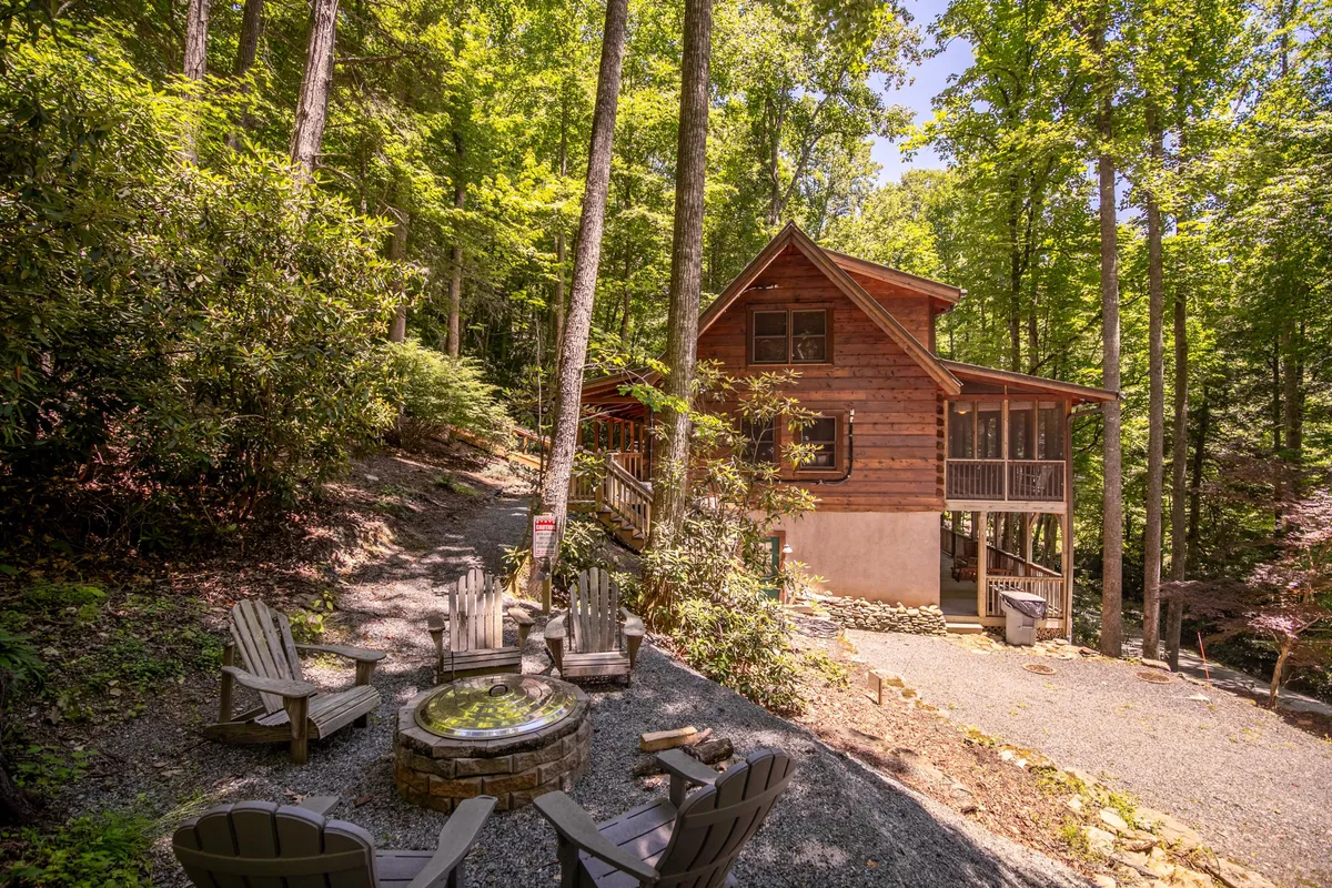 A charming wooden cabin nestled among trees, with an inviting fire pit surrounded by outdoor seating in the foreground. The tranquil setting hints at privacy and the opportunity for cozy evenings under the stars, while the lush greenery and rustic elements evoke a romantic mountain escape.