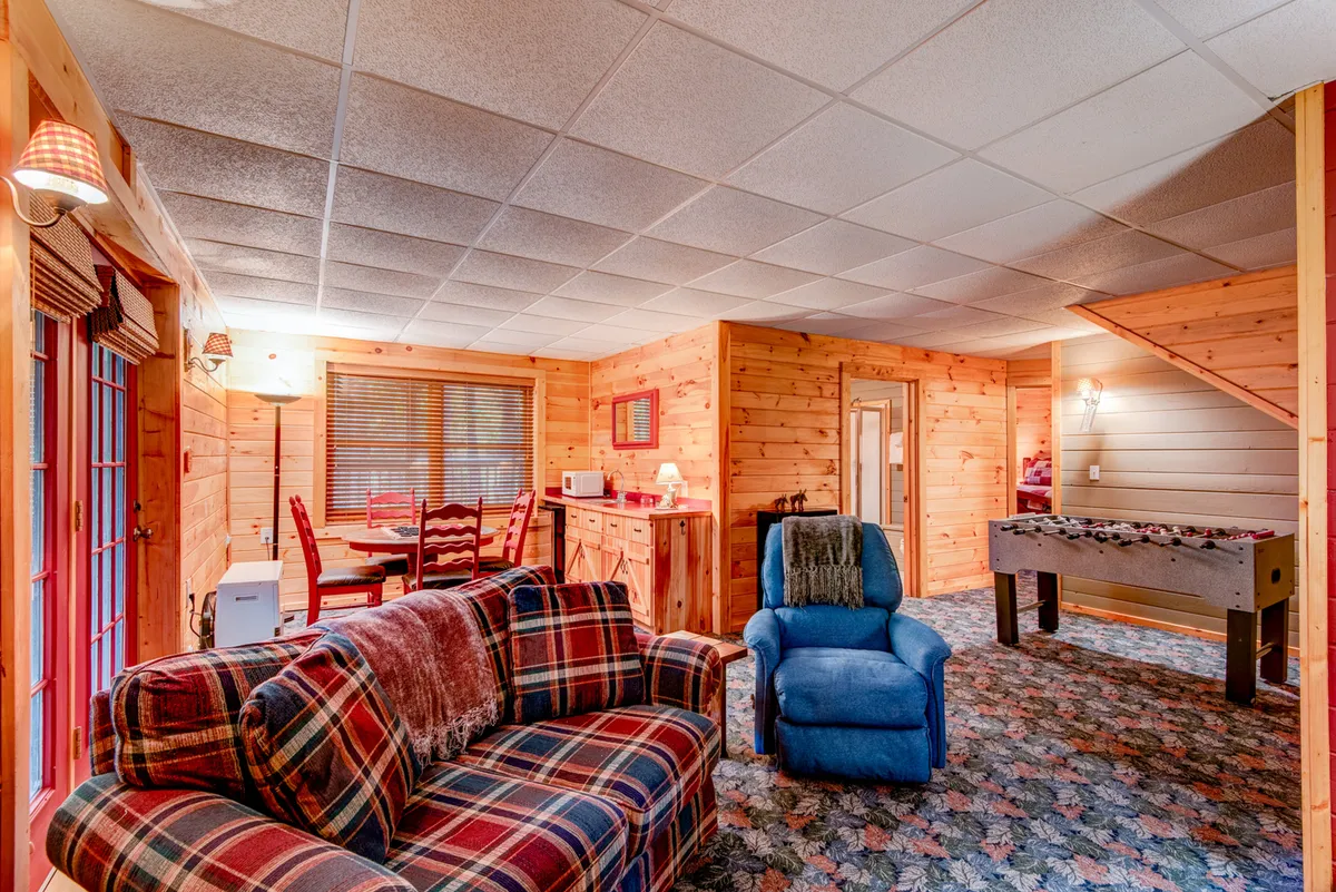 This image shows a relaxed and inviting game room with a plaid sofa, blue recliner, dining table, and most importantly, a foosball table. The arrangement highlights an area designed for both play and comfort, ideal for family gatherings and friendly competitions.