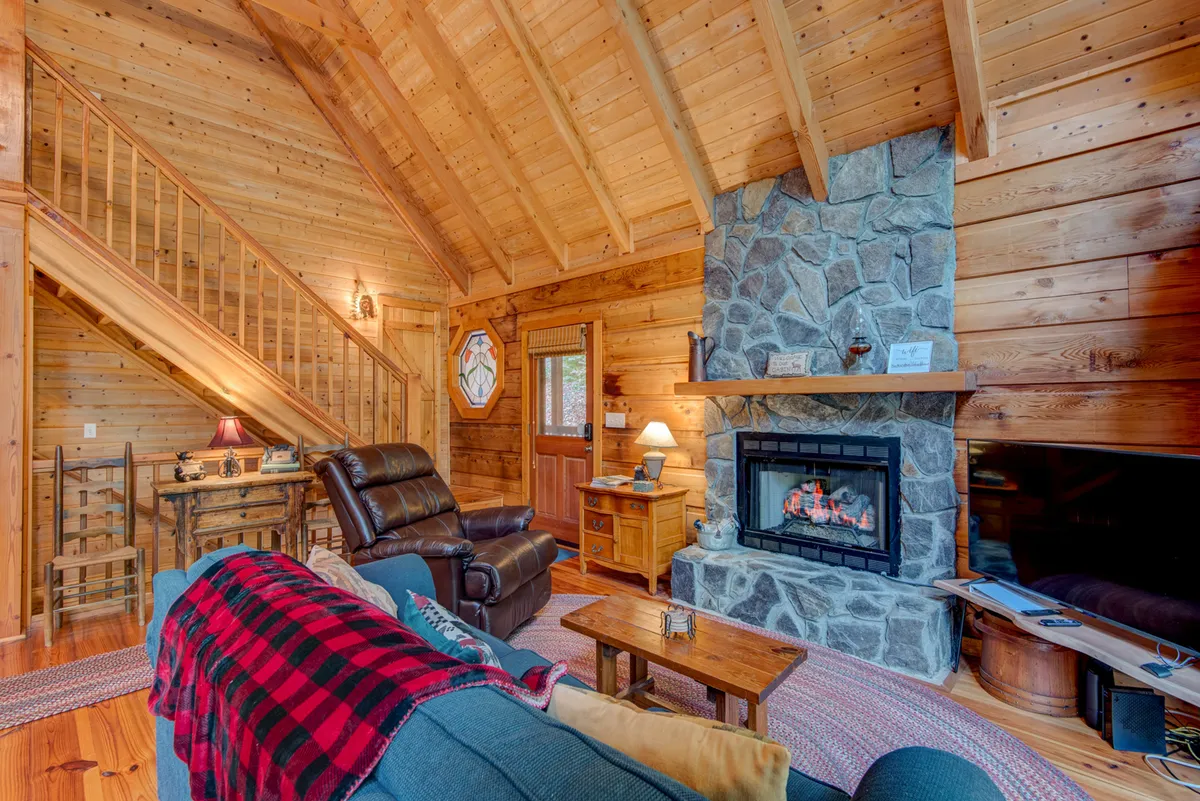 A welcoming living room with a prominent stone fireplace at its center, flanked by cozy, comfortable seating. Exposed wooden beams, staircase, and a decorative lamp add to the rustic, homey charm. The seating area is ideal for gathering, storytelling, or relaxing by the fire on cool evenings.