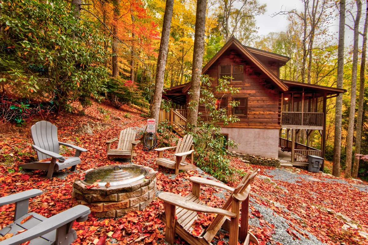 A rustic cabin with a cozy fire pit is framed by Adirondack chairs and brilliant autumn foliage. The seating is arranged for intimate gatherings around the fire, while the vibrant colors and natural setting underscore the beauty of outdoor living and relaxation.